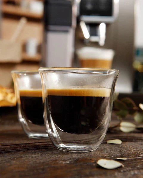 Two clear glass De'Longhi espresso cups filled with fresh espresso on a wooden table with a coffee machine in the background