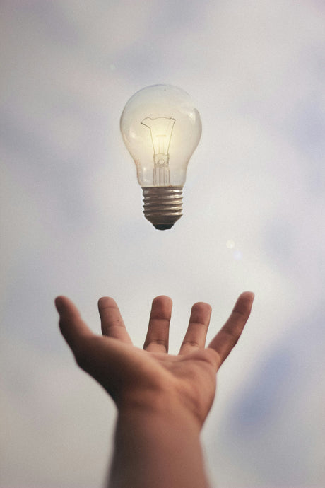 Hand reaching towards a glowing energy efficient light bulb floating against a soft sky background