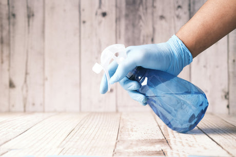 Hand wearing blue glove holding blue spray bottle among cleaning accessories for home on wooden surface