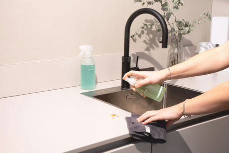 Person using home cleaning products to spray and wipe kitchen countertop near black faucet