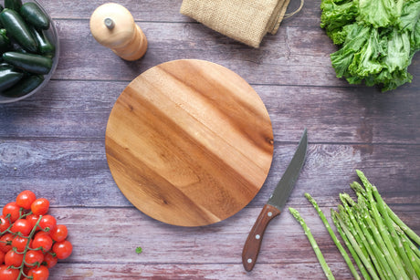 Round wooden chopping board for kitchen with knife surrounded by fresh vegetables on wooden table