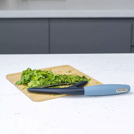 Calitek herb chopper mezzaluna with blue handle on white countertop next to chopped herbs on wooden cutting board