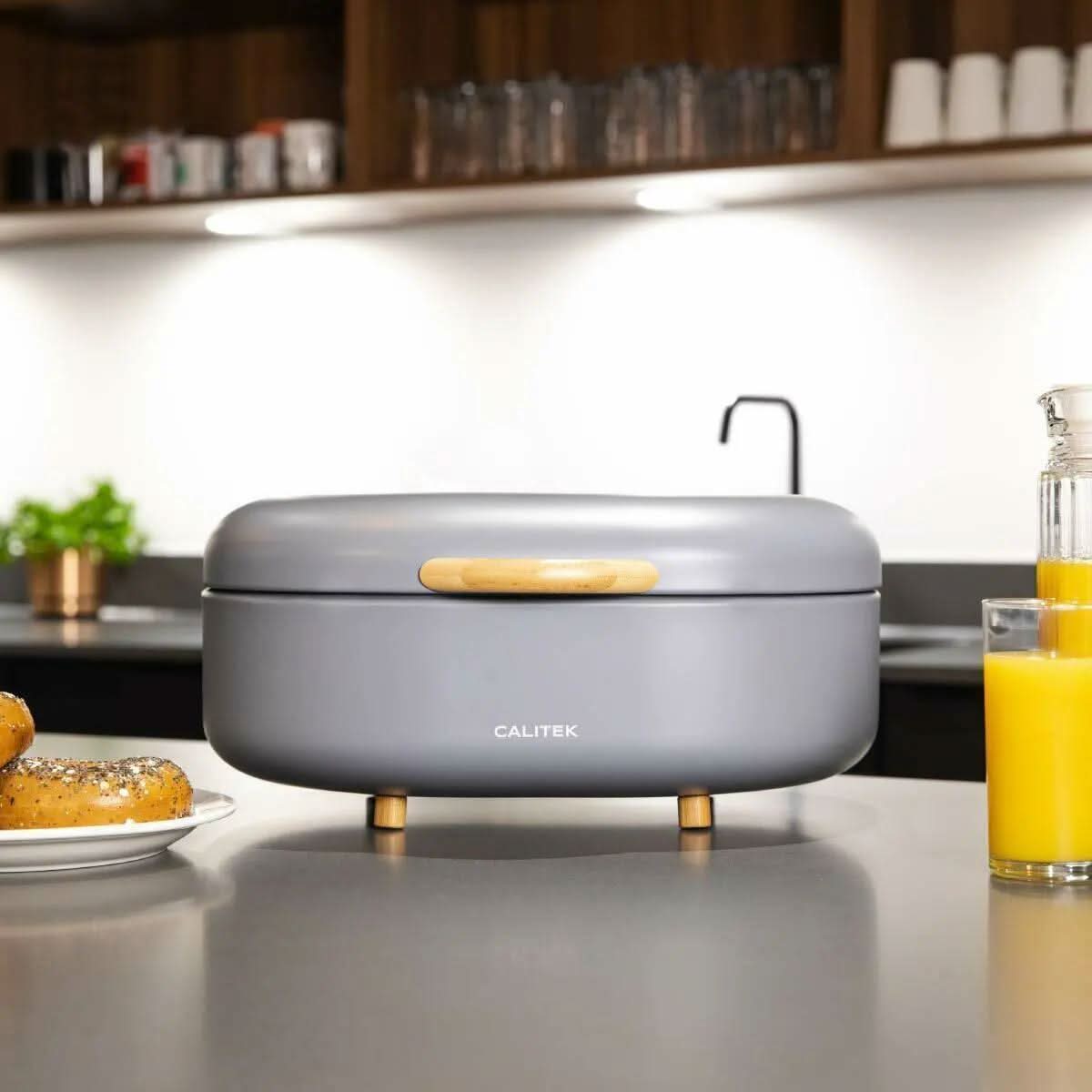 Modern bread bin with bamboo feet in gray on kitchen countertop with orange juice and bagels