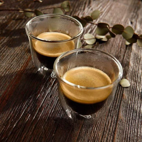 Two DeLonghi double wall espresso glasses filled with espresso on a wooden surface with leaves