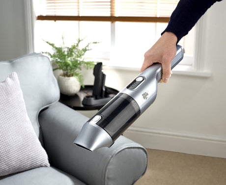 Person using a silver Dirt Devil handheld vacuum to clean a gray sofa in a bright living room