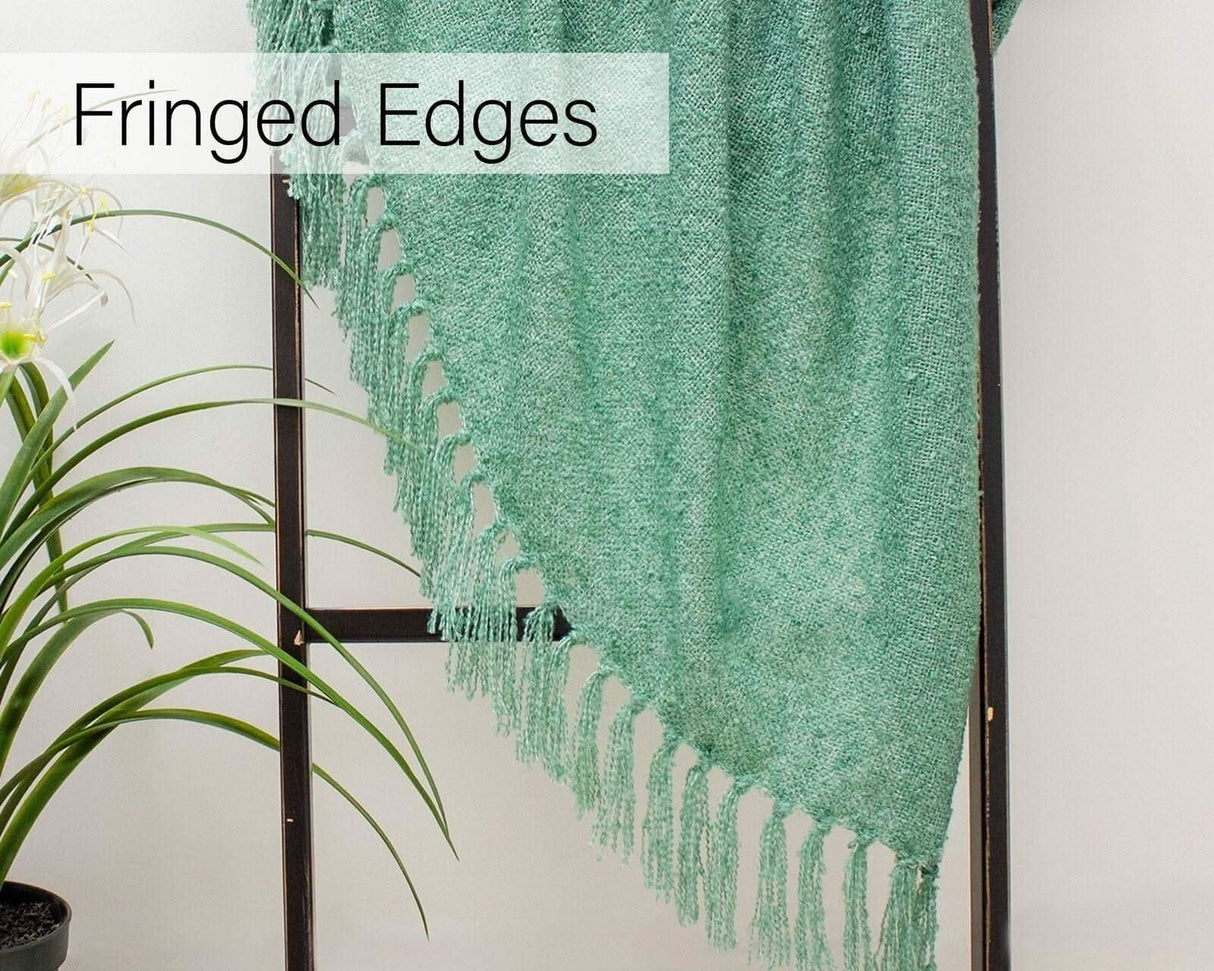 Green loose weave throw blanket with fringed edges hanging on a black ladder next to a green plant