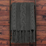 Light grey throw blanket with cable knit pattern and fringed edges displayed on wooden surface