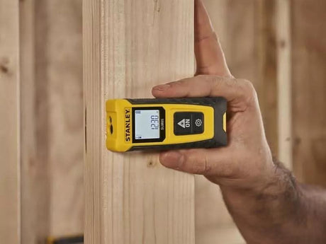Stanley laser distance measure in yellow and black held against wooden plank by hand