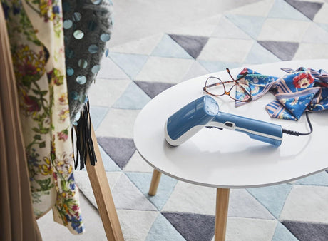Blue Tefal handheld garment steamer on white table with clothes and glasses nearby on patterned rug