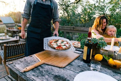 Person wearing heavy duty canvas apron holding a pizza peel with freshly baked pizza outdoors