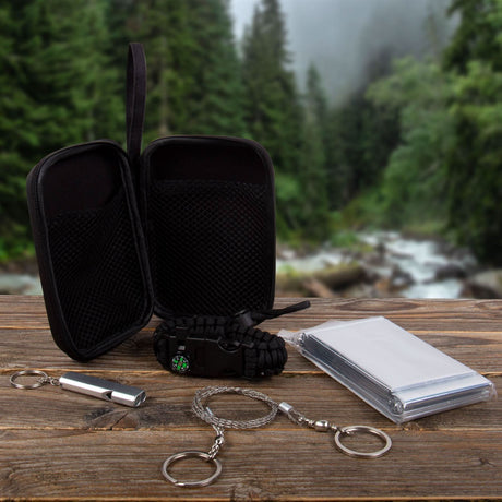 Outdoor survival kit with black carrying case, whistle, paracord bracelet, wire saw, and emergency blanket on wooden surface